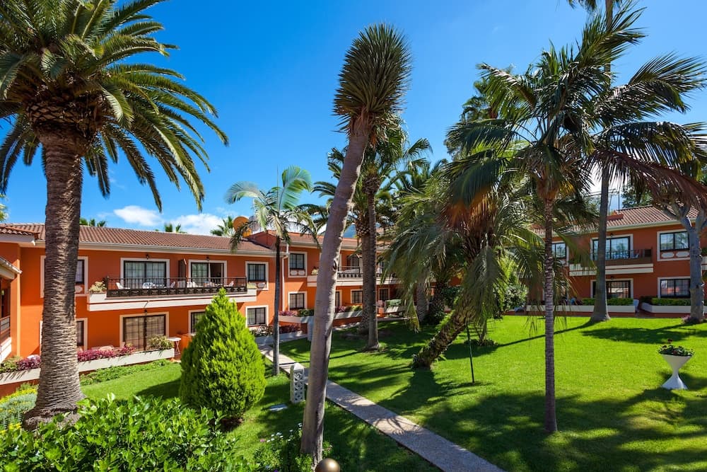 A vibrant orange resort building surrounded by lush green lawns, towering palm trees, and clear blue skies creates an inviting tropical getaway atmosphere ideal for vacation planning.