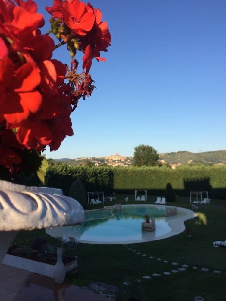 A serene garden scene features vibrant red flowers in the foreground, overlooking a tranquil pool surrounded by lush greenery and distant hills, ideal for a relaxing vacation retreat.
