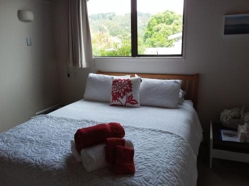 A cozy bedroom with a neatly made bed adorned with red towels and floral pillows overlooks lush greenery through a large window, ideal for travelers seeking a tranquil retreat in a serene natural setting.