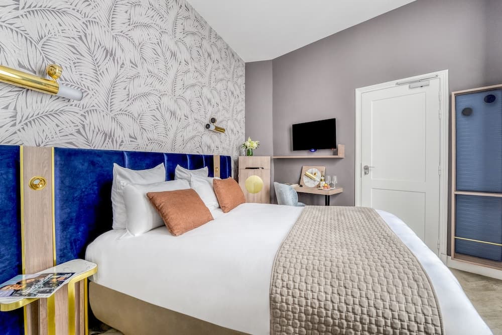 A cozy hotel room features a plush bed with a blue velvet headboard against a leaf-patterned wall, complemented by modern decor and amenities, ideal for travelers seeking comfort and style.