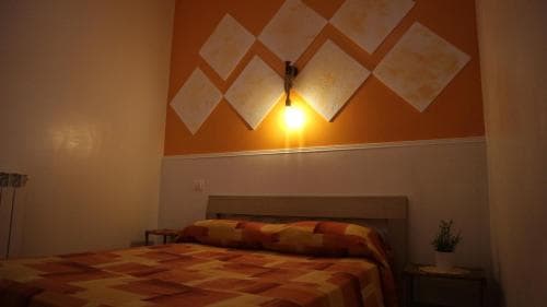 A cozy hotel room features a neatly made bed under warm lighting from a wall-mounted lamp, set against an orange wall with decorative panels, ideal for travelers seeking comfort and simplicity.