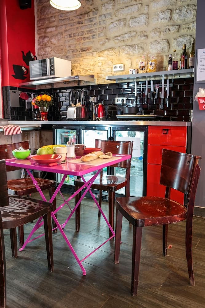 A cozy café corner features a vibrant pink table set with fresh bread and fruit, surrounded by rustic wooden chairs, against a backdrop of a brick wall and modern kitchen appliances, ideal for travelers seeking a charming spot for breakfast or a casual meal.