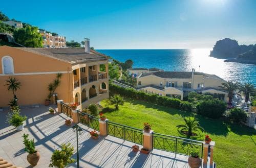 A serene coastal resort features a sunlit terrace adorned with potted plants overlooking lush greenery and the sparkling sea, ideal for travelers seeking relaxation and scenic beauty.