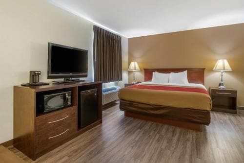 A cozy hotel room features a neatly made bed, wooden furnishings, a flat-screen TV, and warm lighting, ideal for travelers seeking comfort and convenience during their stay.