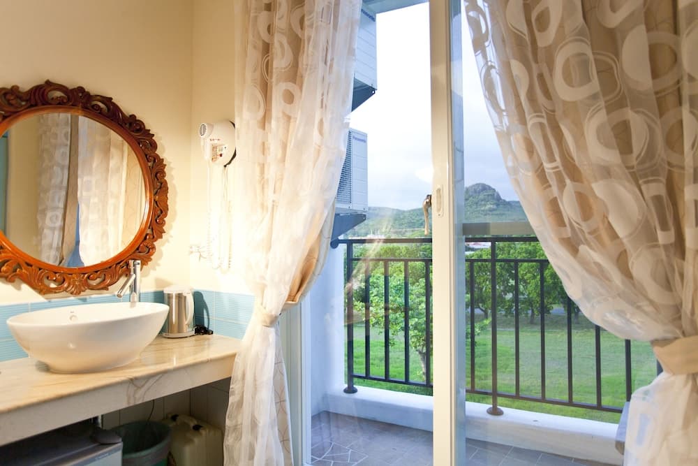 A cozy bathroom with an ornate mirror and sink opens to a serene balcony view of lush greenery, ideal for travelers seeking relaxation in a tranquil setting.