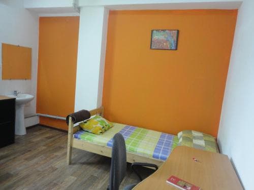 A cozy hostel room with vibrant orange walls, a single bed adorned with colorful bedding, a desk with a chair, and a small sink area, ideal for budget travelers seeking a comfortable and affordable stay.