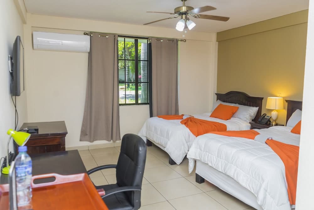 A cozy hotel room with two neatly made beds adorned in orange accents, a desk with a chair, and a view of greenery through the window, ideal for travelers seeking comfort and convenience.