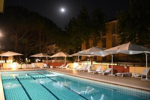 A serene night-time view of an outdoor hotel pool area illuminated by the moonlight, featuring lounge chairs under umbrellas, surrounded by trees, ideal for travelers seeking relaxation and nighttime ambiance.