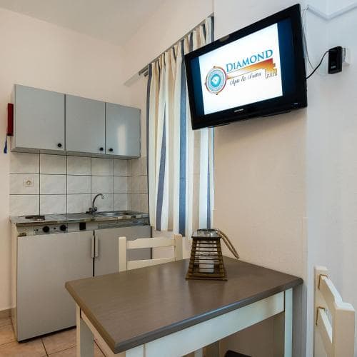 A cozy studio apartment features a compact kitchenette, dining table, and mounted TV displaying a logo, ideal for travelers seeking a functional and comfortable stay in an urban setting.