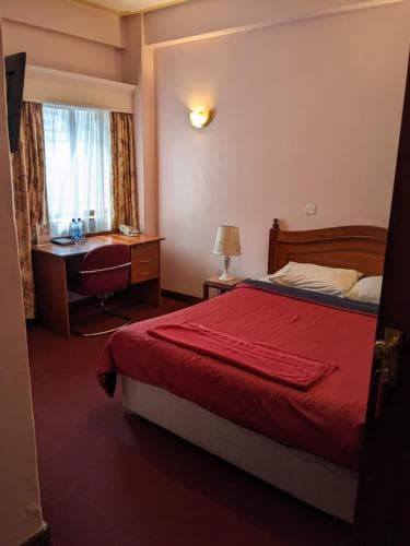 A cozy hotel room features a neatly made bed with red bedding, a wooden desk by the window with curtains, and warm lighting, ideal for travelers seeking comfort and functionality.