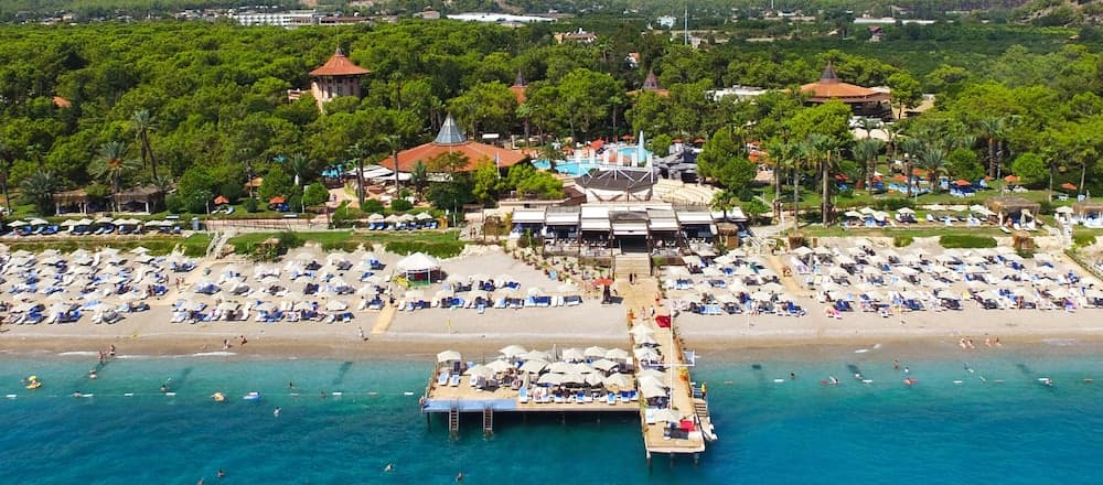 An aerial view captures a vibrant beach resort with turquoise waters, rows of sun loungers under white umbrellas, lush greenery, and a pier extending into the sea, ideal for travelers seeking relaxation and tropical escapism.