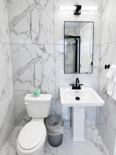 A pristine bathroom with white marble walls exudes elegance, featuring a sleek sink, modern toilet, and reflective mirror, ideal for travelers seeking luxury accommodations.