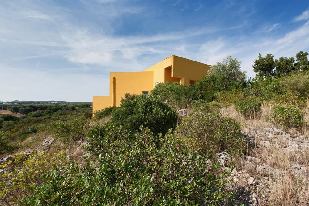 A vibrant yellow modern house stands amidst lush greenery on a rocky hillside under a serene blue sky, ideal for travelers seeking a tranquil retreat in nature.
