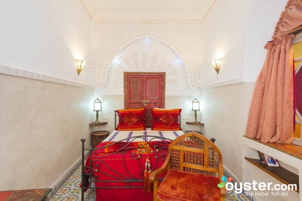 A cozy, ornately decorated bedroom with a red bedspread, intricate wall designs, and warm lighting creates an inviting atmosphere ideal for travelers seeking a luxurious and culturally rich accommodation experience.
