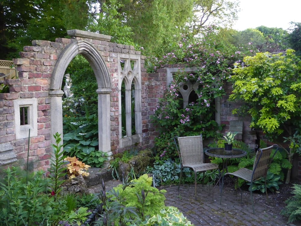 A serene garden patio features a rustic brick archway adorned with lush greenery and pink flowers, creating an inviting atmosphere perfect for a tranquil retreat or romantic getaway.