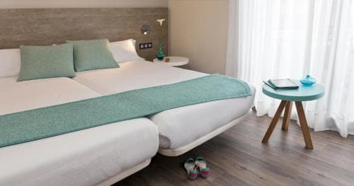 A cozy hotel room with a neatly made bed adorned in teal accents, a small wooden side table with a book and vase, and soft natural light filtering through sheer curtains, ideal for travelers seeking comfort and relaxation.