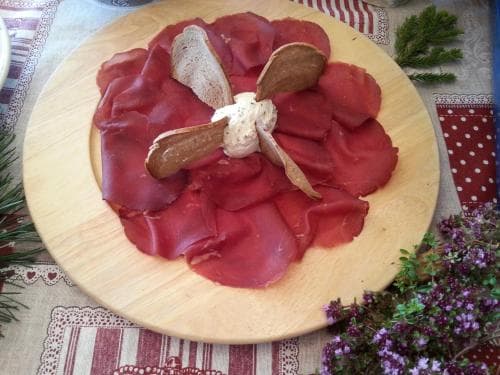 A rustic wooden platter adorned with thinly sliced cured meat, garlic cloves, and herbs creates an inviting atmosphere on a patterned tablecloth, perfect for a cozy culinary exploration or food photography session.