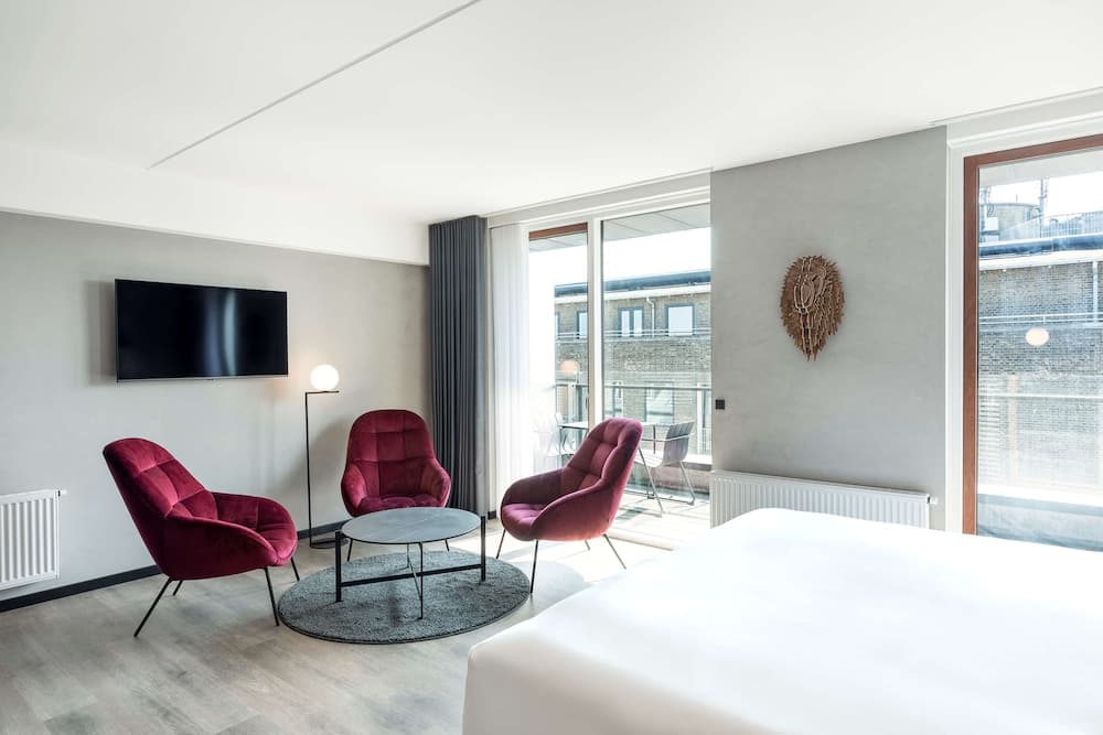 A modern hotel room features plush red armchairs, a sleek coffee table, and large windows with urban views, creating a cozy yet stylish atmosphere ideal for travelers seeking comfort and contemporary design.