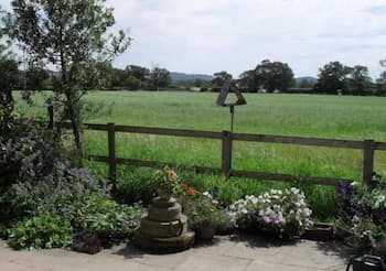 A serene garden path lined with vibrant flowers and a stone stack leads to a wooden fence overlooking lush green fields, ideal for tranquil countryside retreats.