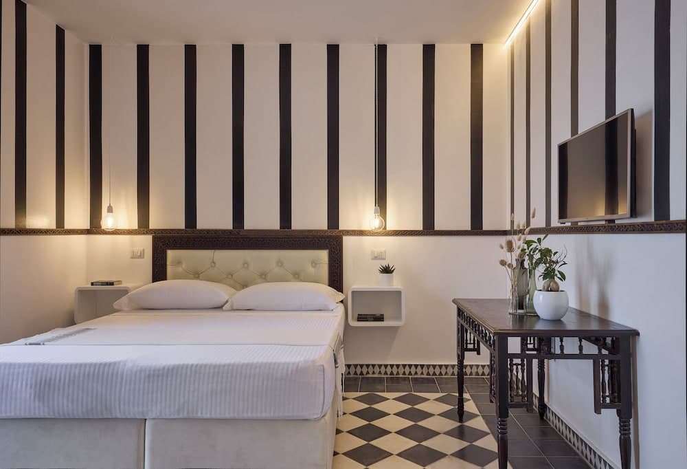 A cozy hotel room features a stylish black-and-white striped wall, a neatly made bed with white linens, ambient lighting, and a checkered floor, ideal for travelers seeking comfort and modern aesthetics.