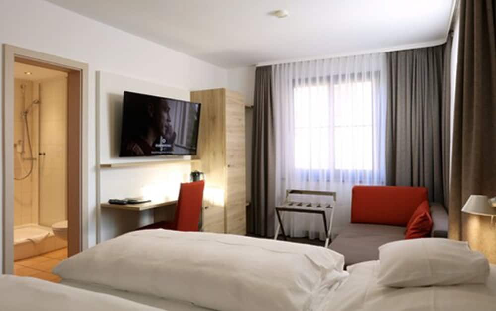 A cozy hotel room features a neatly made bed, a modern desk setup with a mounted TV, a comfortable seating area by the window, and an adjacent bathroom, ideal for travelers seeking comfort and convenience.