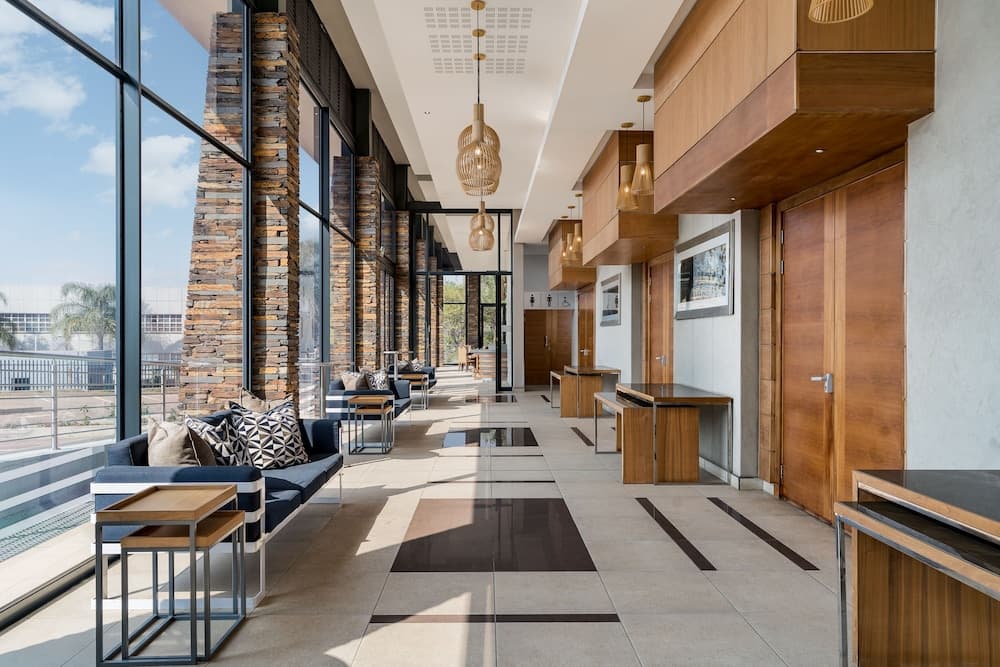 A modern hotel lobby features sleek seating areas with patterned cushions, wooden tables, and large windows offering a serene outdoor view, ideal for travelers seeking a relaxing and stylish atmosphere.