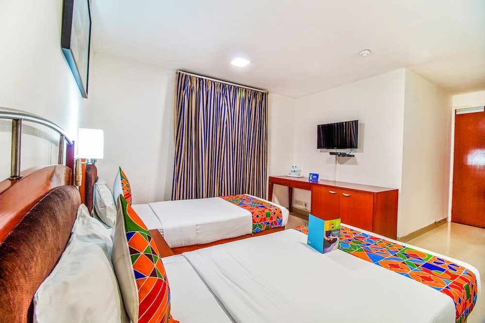 A cozy hotel room features two neatly made beds adorned with vibrant geometric-patterned throws, a wooden desk with a mounted TV, colorful striped curtains, and warm lighting, ideal for travelers seeking comfort and convenience.