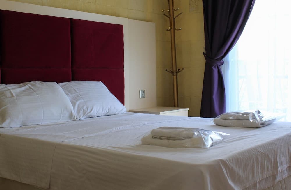 A cozy hotel room features a neatly made bed with white linens, a burgundy headboard, purple curtains, and a sunny balcony view, ideal for travelers seeking comfort and relaxation.
