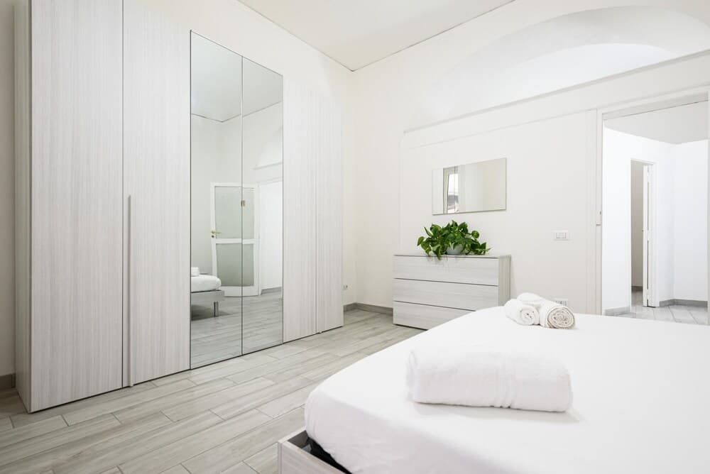 A serene minimalist bedroom with white furnishings, mirrored closet doors, and a fresh green plant exudes calmness, ideal for travelers seeking a peaceful retreat.