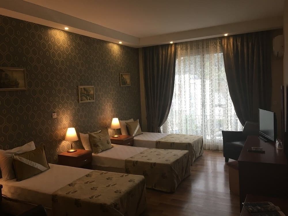 A cozy hotel room with three neatly arranged beds, patterned wallpaper, soft lighting, and a large window with sheer curtains creates a welcoming atmosphere ideal for family travelers seeking comfort and relaxation.