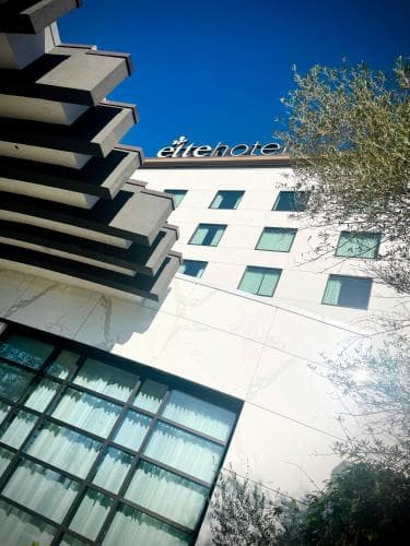 A modern hotel facade with "elitehotel" signage stands under a clear blue sky, framed by lush greenery, ideal for travelers seeking urban accommodation with a touch of nature.