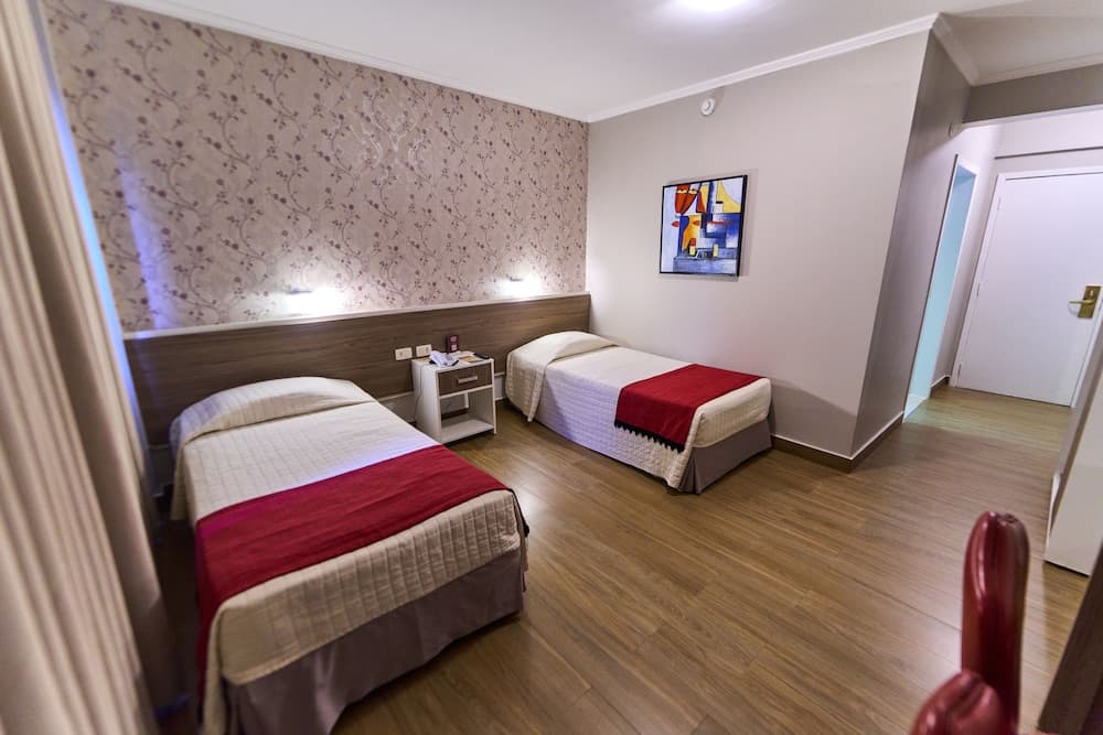 A cozy hotel room features two neatly made beds with red accents, floral wallpaper, modern artwork, and wooden flooring, ideal for travelers seeking comfort and style.