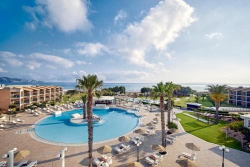 A vibrant resort scene featuring a large pool surrounded by palm trees, lounge chairs, and a picturesque ocean view under a partly cloudy sky, ideal for a relaxing beach vacation getaway.