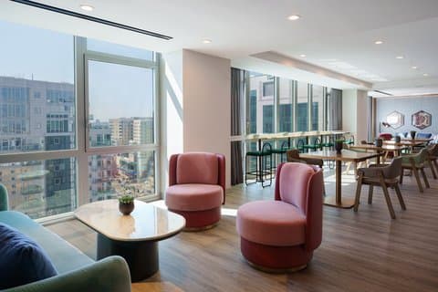 A modern lounge area with plush pink chairs and large windows overlooking an urban skyline creates a cozy yet vibrant atmosphere in the Renaissance New York Flushing Hotel at Tangram, ideal for travelers seeking comfort and style during their stay.