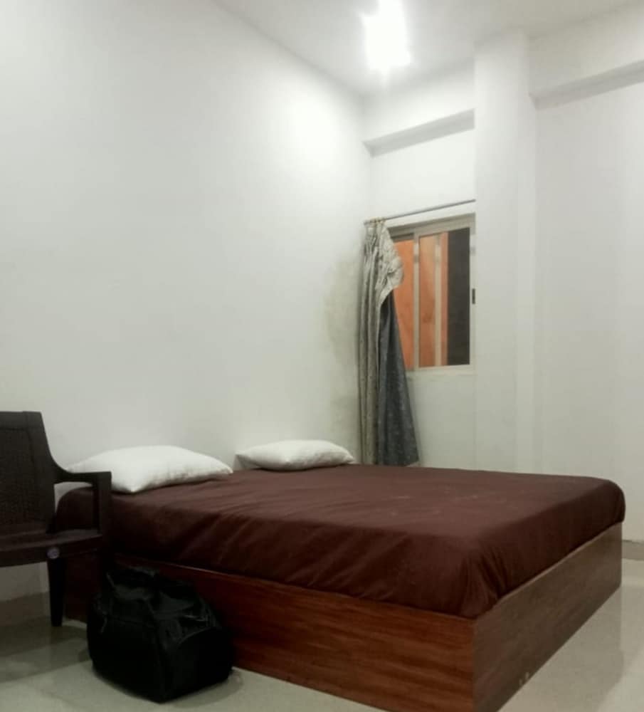 A minimalist hotel room features a neatly made bed with a brown cover, white pillows, a simple chair, and a window with curtains, offering a cozy and budget-friendly accommodation option for travelers seeking comfort in a clean space.