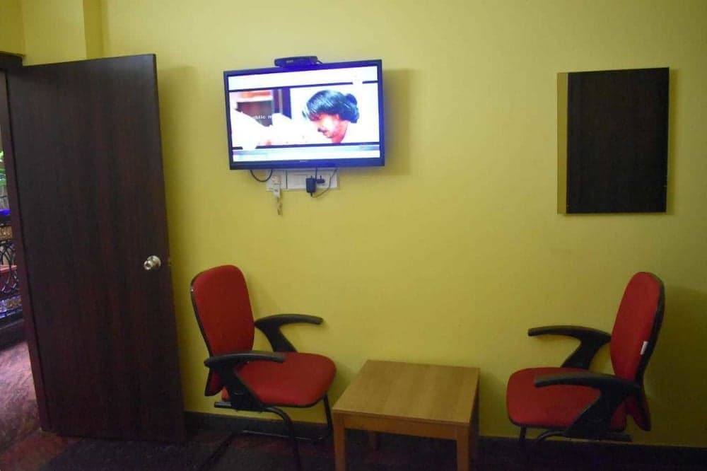 A cozy waiting area features vibrant yellow walls, red chairs, a small wooden table, and a mounted TV displaying a news program, ideal for travelers seeking a comfortable spot to relax and stay informed.