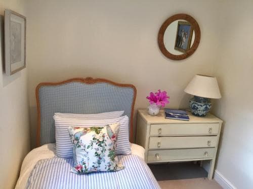 A cozy bedroom corner features a charming single bed with floral pillows, a vintage dresser adorned with a blue lamp and pink flowers, and a round mirror reflecting a serene atmosphere, ideal for travelers seeking a quaint and comfortable retreat.