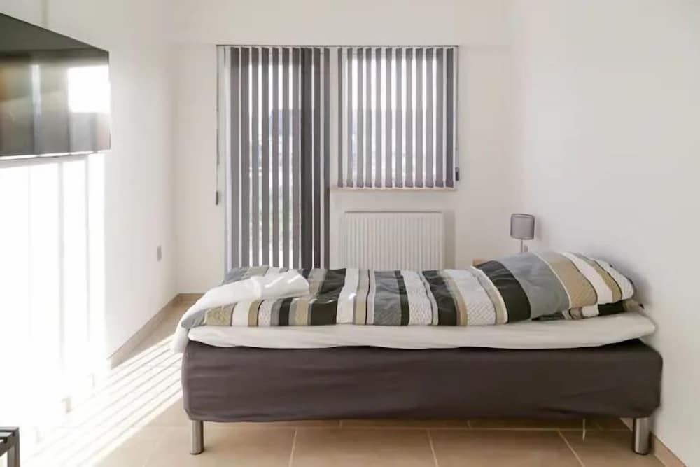 A minimalist bedroom with a neatly made bed, vertical blinds on the window, and soft lighting creates a serene atmosphere ideal for travelers seeking a cozy and tranquil accommodation.