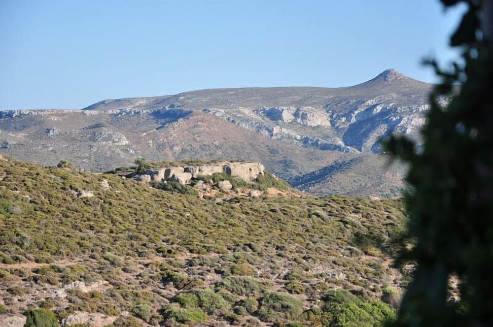 A serene landscape featuring ancient stone ruins atop a lush hillside under a clear blue sky, ideal for history enthusiasts exploring Mediterranean archaeological sites.