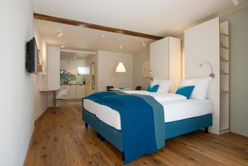 A cozy hotel suite features a modern bedroom with a blue-accented bed, wooden floors, and an open-plan kitchen area, ideal for travelers seeking comfort and convenience in a stylish urban retreat.
