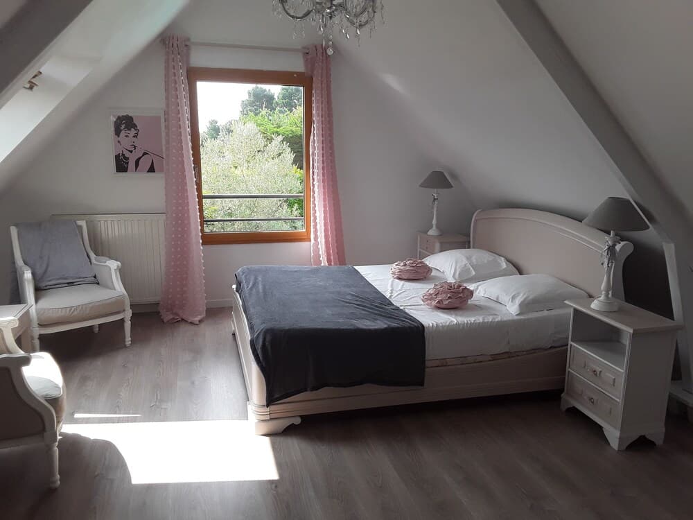A cozy attic bedroom with sloped ceilings, pink curtains, and a serene view of greenery through the window, featuring elegant white furniture, a chandelier, and a stylish travel accommodation vibe.