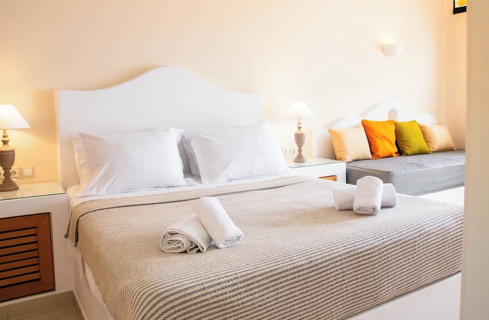 A cozy hotel bedroom features a neatly made bed with white linens, rolled towels, a colorful cushioned bench, and soft lighting, ideal for travelers seeking comfort and relaxation.
