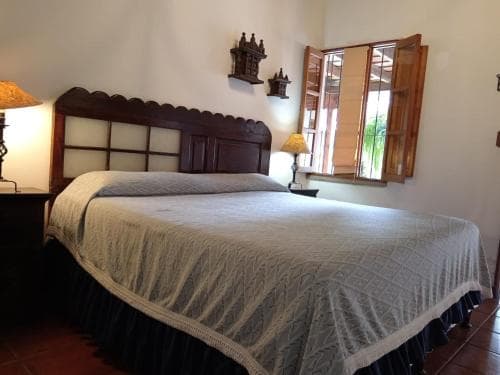 A cozy bedroom with a neatly made bed, wooden furnishings, and open shutters revealing greenery outside, ideal for travelers seeking a tranquil retreat in a rustic setting.
