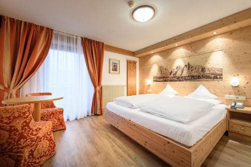 A cozy hotel room with wooden walls, a neatly made bed, vibrant orange curtains, and mountain artwork exudes warmth and comfort, ideal for travelers seeking relaxation in an alpine setting.