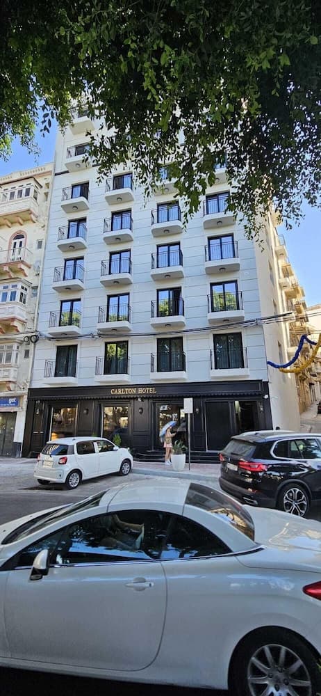 A serene urban scene features the Carlton Hotel, a white building with balconies framed by lush greenery, parked cars, and a pedestrian, ideal for travelers seeking accommodation in a bustling city center.