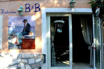 A charming B&B entrance features a rustic stone wall, a vibrant mural of a sailor by the sea, and inviting curtains, perfect for travelers seeking cozy seaside accommodations.