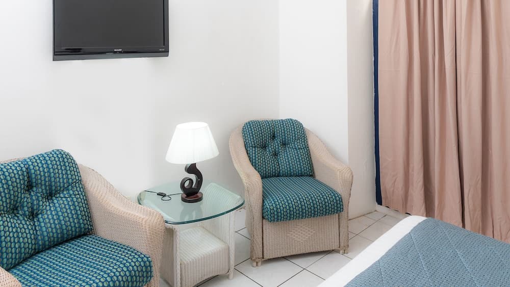 A cozy hotel room corner features two wicker chairs with blue patterned cushions, a glass side table with a modern lamp, and a wall-mounted TV, ideal for travelers seeking comfort and relaxation.