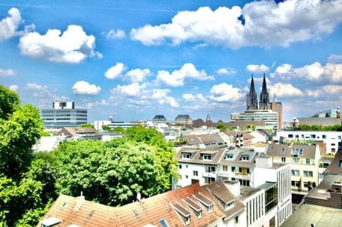 A vibrant cityscape featuring a prominent cathedral under a bright blue sky dotted with fluffy clouds, surrounded by lush greenery and modern buildings, ideal for urban exploration and cultural tourism.