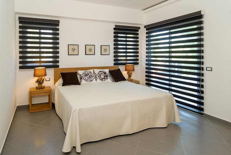 A serene bedroom at Aligio Playa Poppy in Las Terrenas, Dominican Republic, features a cozy bed adorned with patterned pillows, wooden nightstands with lamps, and large windows with striped blinds, ideal for a tranquil vacation retreat.
