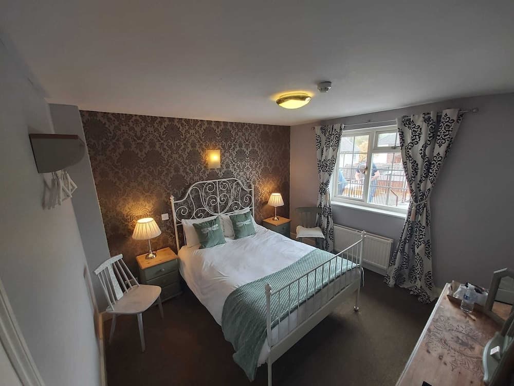 A cozy bedroom with a white metal bed, patterned wallpaper, and a window with decorative curtains offers a serene retreat for travelers seeking comfort and relaxation.